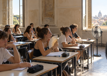 Studenti seduti in un’aula scolastica con finestre aperte e ventilatori accesi.