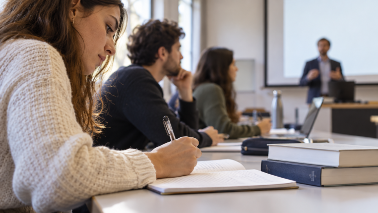 Una studentessa prende appunti durante una lezione universitaria con altri partecipanti