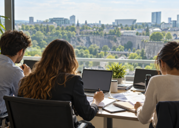 Tre giovani di spalle lavorano su documenti e computer in un ufficio luminoso.