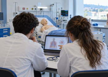 Due giovani in camice studiano al computer in un laboratorio di simulazione sanitaria.