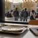 mmagine simbolica: vassoio e quaderno in una mensa, con studenti di spalle nel cortile universitario.
